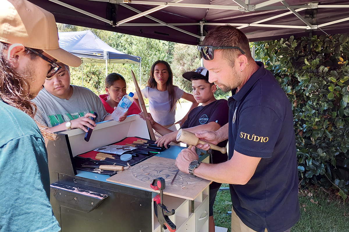 Five people watch a man doing whakairo (carving).